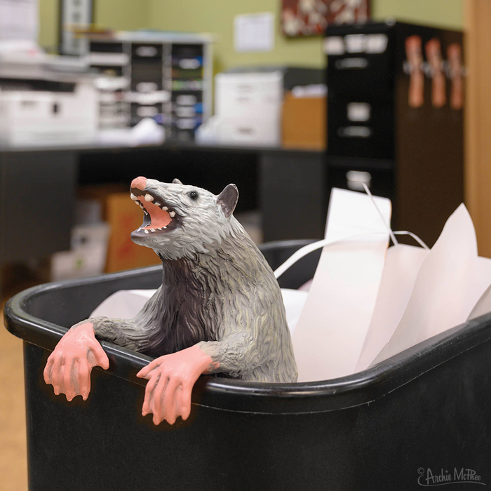 In an office setting, an Office Possum, crafted from stuffed latex and featuring posable paws, peeks out of a trash bin filled with paper.