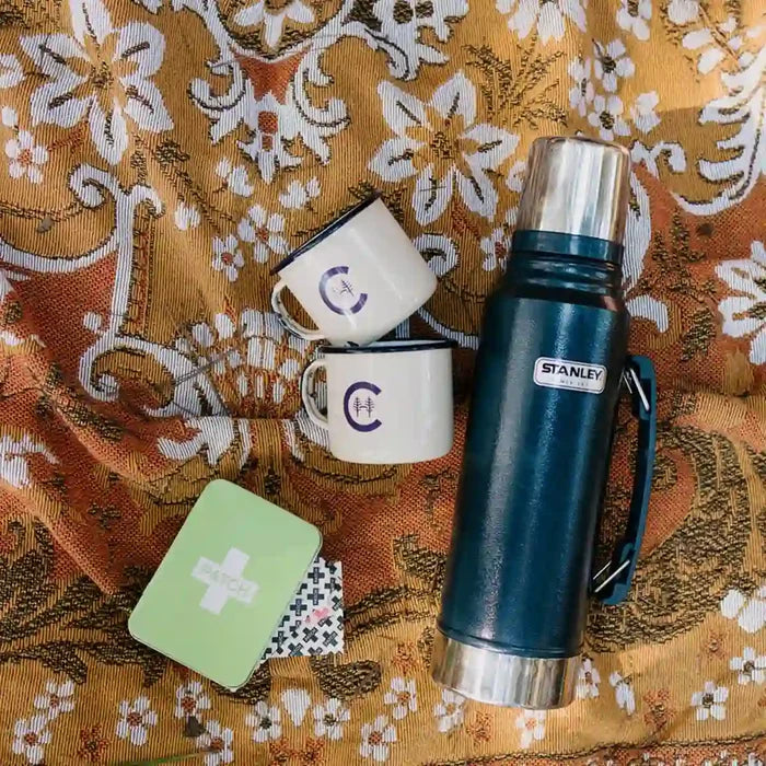 Blue Stanley thermos with two white mugs and a green bandage tin on a floral picnic blanket.