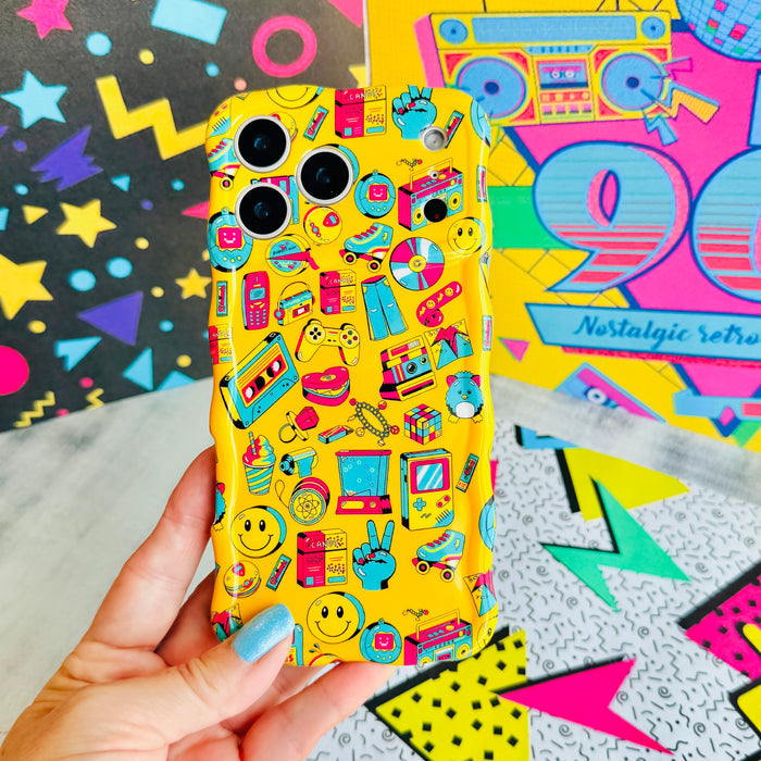 Hand holding the Oops, It's the 90's Wavy Luxe MagSafe iPhone case, showing the yellow print packed with smiley faces, boom boxes, roller skates and retro stickers.