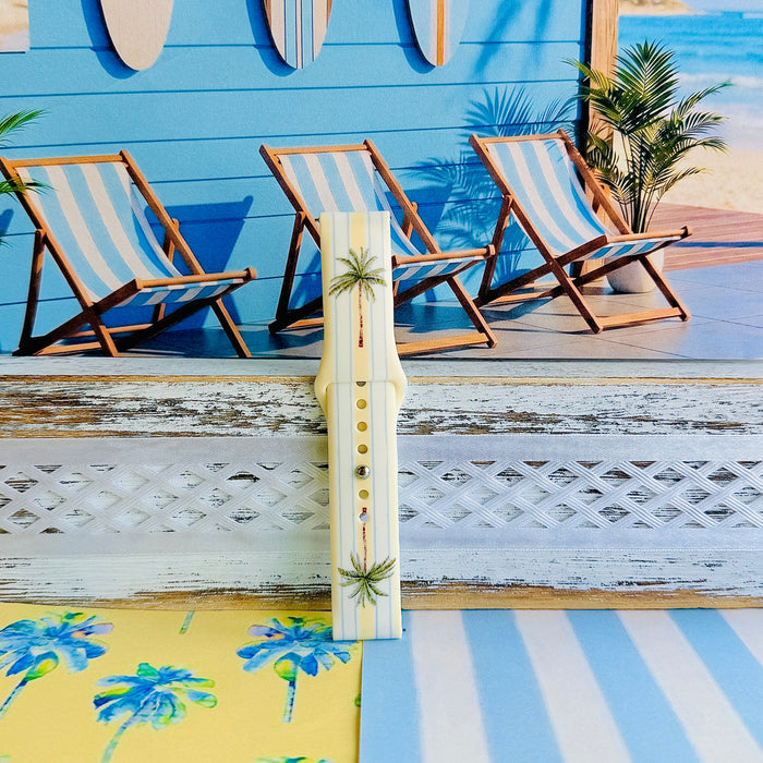 Palm Shore Club silicone Samsung Watch band with hand-painted palm trees over soft blue and yellow cabana stripes on an ivory base, shown displayed and worn on Apple Watch against a coastal beach club backdrop.