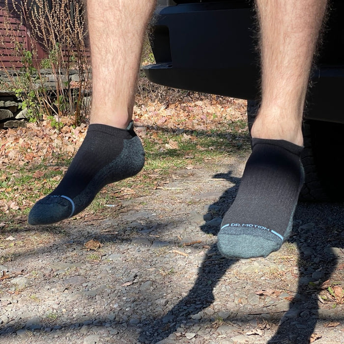 Ankle compression socks in black and gray worn on a person's feet, with a gravel surface and autumn leaves in the background.