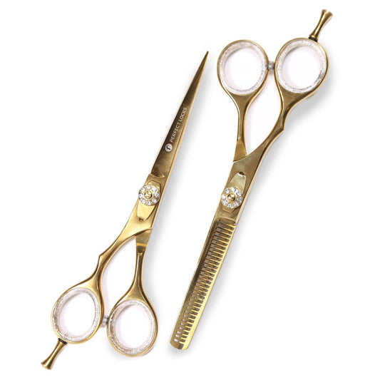 Two gold-colored hairdressing scissors with white finger rings, one with a comb-like thinning blade, lie side by side on a white background.