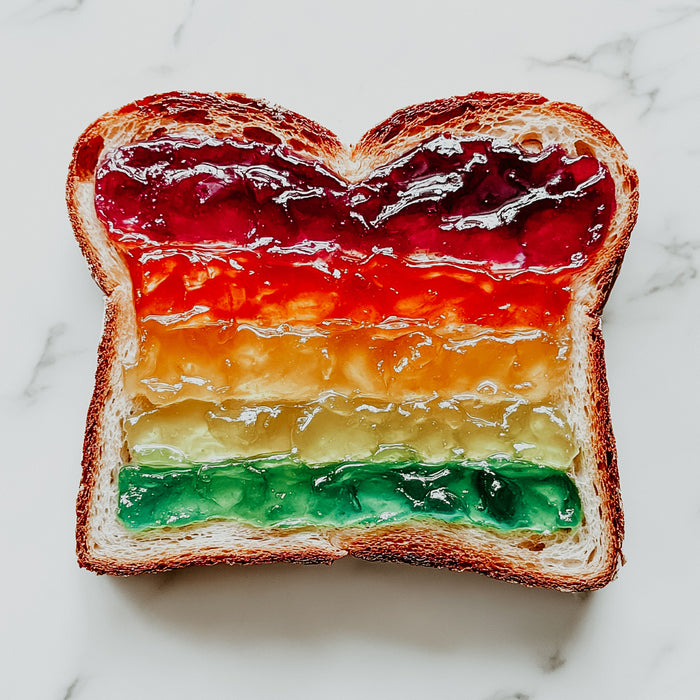 A slice of bread with multicolored layers of jam resembling a rainbow on a white marble surface.