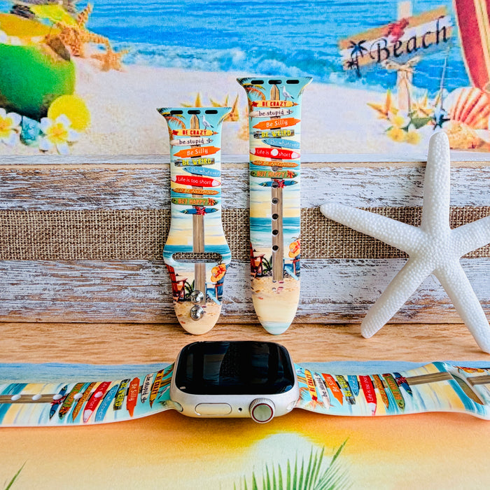 Seaside State of Mind silicone Apple Watch band with colorful surfboard signpost on a beach scene and "Be Crazy, Be Silly, Be Weird—life is too short to be anything but happy" message, shown displayed and worn on Apple Watch against a beach backdrop.