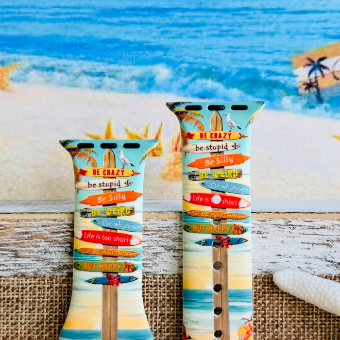 Seaside State of Mind silicone Apple Watch band with colorful surfboard signpost on a beach scene and "Be Crazy, Be Silly, Be Weird—life is too short to be anything but happy" message, shown displayed and worn on Apple Watch against a beach backdrop.