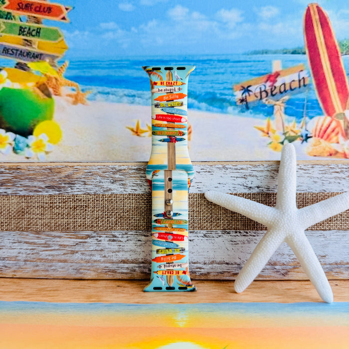 Seaside State of Mind silicone Apple Watch band with colorful surfboard signpost on a beach scene and "Be Crazy, Be Silly, Be Weird—life is too short to be anything but happy" message, shown displayed and worn on Apple Watch against a beach backdrop.