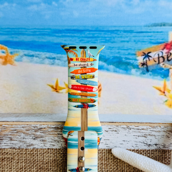 Seaside State of Mind silicone Apple Watch band with colorful surfboard signpost on a beach scene and "Be Crazy, Be Silly, Be Weird—life is too short to be anything but happy" message, shown displayed and worn on Apple Watch against a beach backdrop.