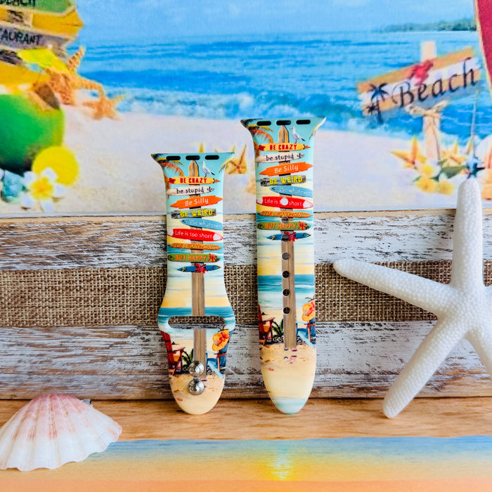 Seaside State of Mind silicone Apple Watch band with colorful surfboard signpost on a beach scene and "Be Crazy, Be Silly, Be Weird—life is too short to be anything but happy" message, shown displayed and worn on Apple Watch against a beach backdrop.
