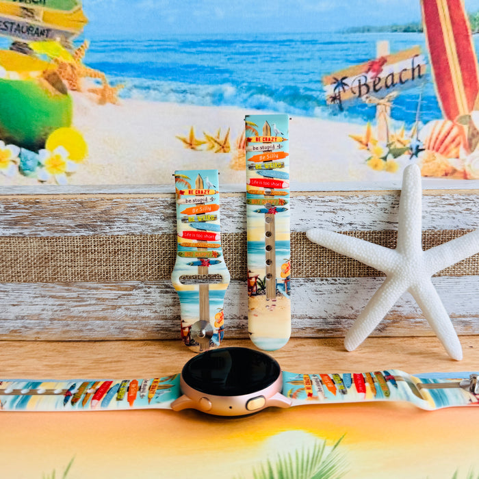 Seaside State of Mind silicone Samsung Watch band with colorful surfboard signpost on a beach scene and "Be Crazy, Be Silly, Be Weird—life is too short to be anything but happy" message, shown displayed and worn on Apple Watch against a beach backdrop.