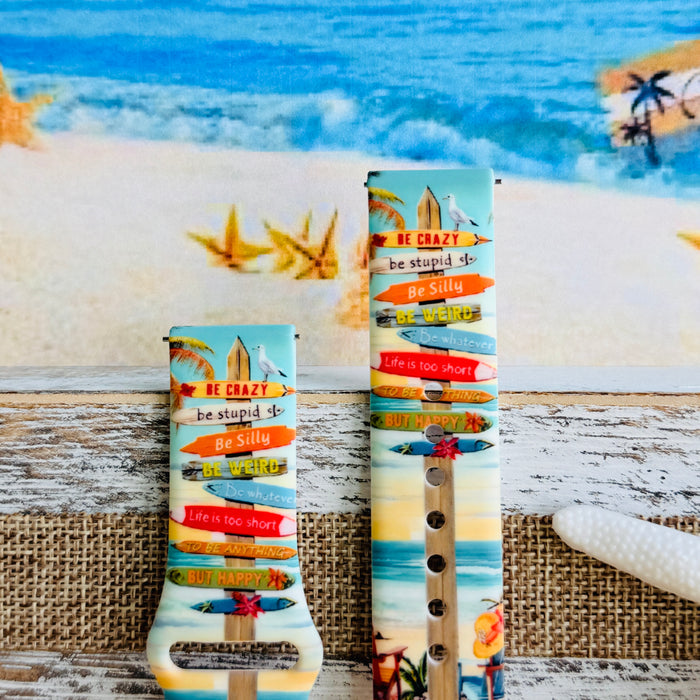 Seaside State of Mind silicone Samsung Watch band with colorful surfboard signpost on a beach scene and "Be Crazy, Be Silly, Be Weird—life is too short to be anything but happy" message, shown displayed and worn on Apple Watch against a beach backdrop.