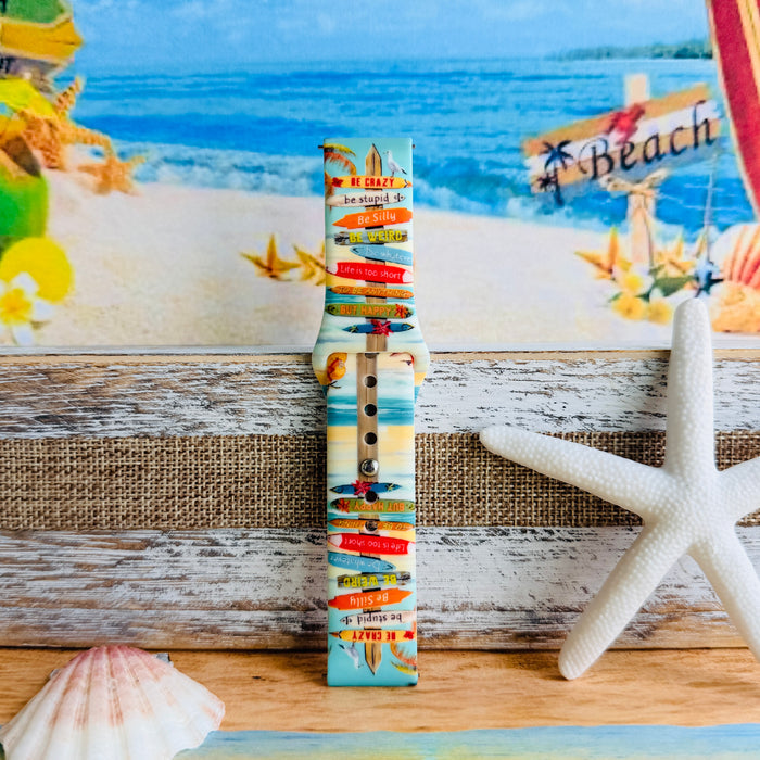 Seaside State of Mind silicone Samsung Watch band with colorful surfboard signpost on a beach scene and "Be Crazy, Be Silly, Be Weird—life is too short to be anything but happy" message, shown displayed and worn on Apple Watch against a beach backdrop.