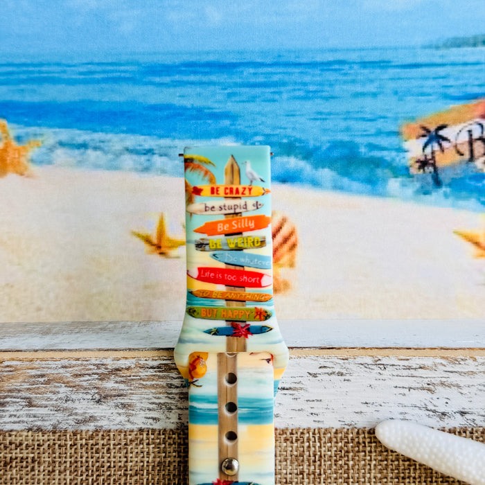 Seaside State of Mind silicone Samsung Watch band with colorful surfboard signpost on a beach scene and "Be Crazy, Be Silly, Be Weird—life is too short to be anything but happy" message, shown displayed and worn on Apple Watch against a beach backdrop.