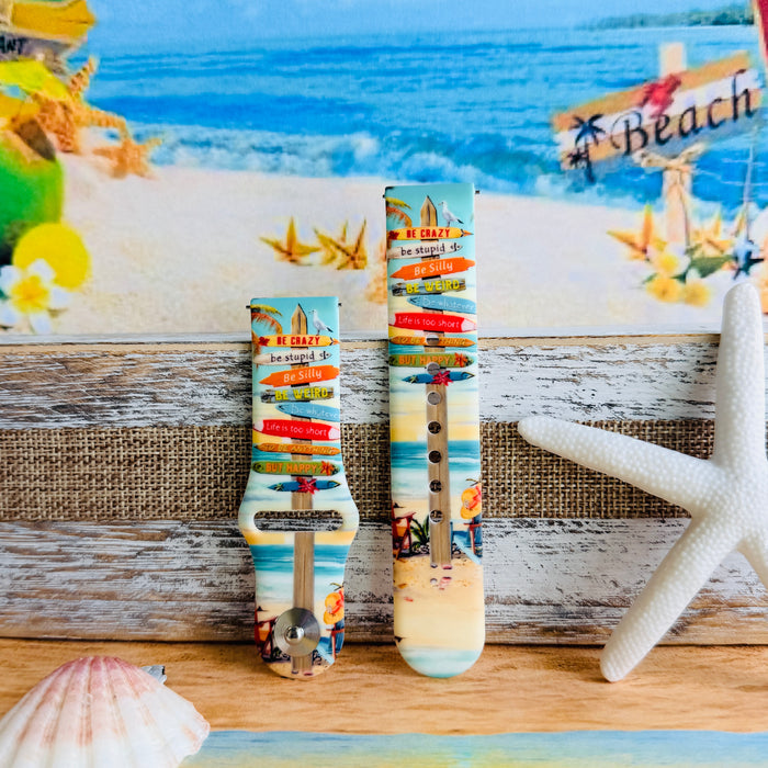 Seaside State of Mind silicone Samsung Watch band with colorful surfboard signpost on a beach scene and "Be Crazy, Be Silly, Be Weird—life is too short to be anything but happy" message, shown displayed and worn on Apple Watch against a beach backdrop.