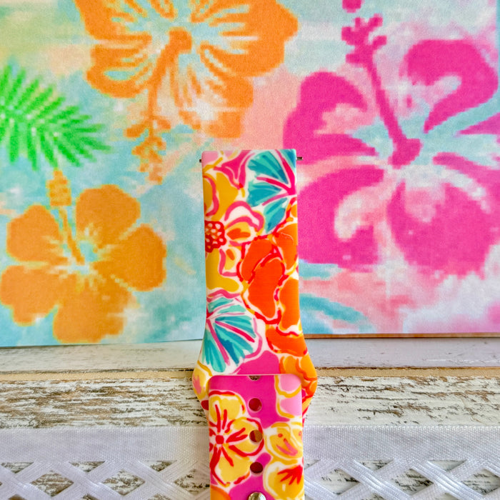 Sunset Hibiscus silicone Samsung Watch band with vibrant pink, orange, yellow, and teal tropical hibiscus floral print, shown displayed and worn on Apple Watch against a colorful tropical floral backdrop.